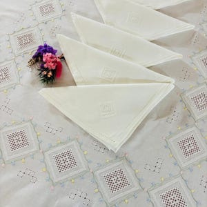 Op de afbeelding: Een crèmekleurig tafelkleed met ingewikkeld wit ajourborduurwerk in ruitpatronen met kleine pastelkleurige bloemaccenten. Verschillende gevouwen crèmekleurige servetten liggen erop gestapeld, elk met een centraal vierkant borduurdetail.