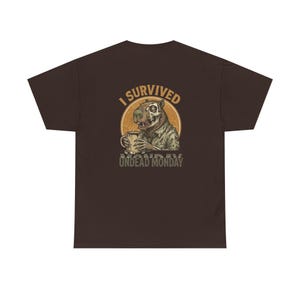 Könnte beinhalten: Dunkelbraunes T-Shirt mit einer Grafik einer zombieartigen Kreatur, die eine Tasse hält. Der Text "I SURVIVED UNDEAD MONDAY" ist oberhalb und unterhalb der Illustration in einem Kreis aufgedruckt.