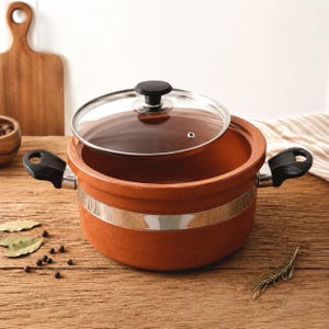 May include: A terracotta cooking pot with a clear glass lid and black handle. The pot has a silver band around the middle and black handles on each side. The lid has a black knob. The pot is sitting on a wooden surface.