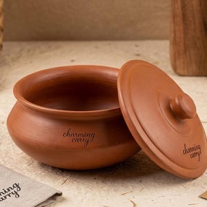 Könnte beinhalten: Ein terracottafarbener Tontopf mit passendem Deckel, leicht geöffnet. Der Topf hat eine abgerundete Form mit einer weiten Öffnung. Der Deckel hat einen kleinen Knopf. Die Worte "charming carry" sind auf Topf und Deckel aufgedruckt.