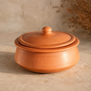 Può includere: Una pentola rotonda in terracotta con coperchio abbinato. Il recipiente presenta una finitura liscia in argilla color marrone rossastro, una base larga che si restringe leggermente verso l'alto e un piccolo pomello centrale sul coperchio.