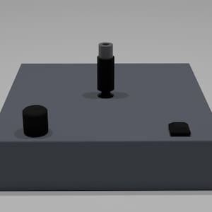 May include: A gray rectangular block with three black objects on top. One is cylindrical, one is a small square, and the other is a taller, cylindrical object with a gray top. The objects are evenly spaced.