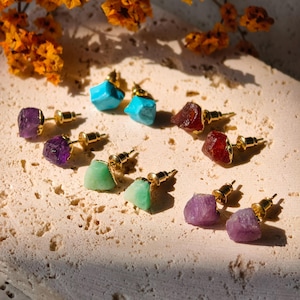 May include: A collection of stud earrings featuring raw, uncut gemstones in various colors including purple, turquoise, red, and light green. Each stone is set on a gold-toned metal post with a secure backing, showcasing natural textures.