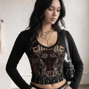 May include: Black long-sleeved crop top with a V-neck and a graphic design. The design includes ornate details and the word "HIPETAL". The top is worn with a silver belt and a black handbag.