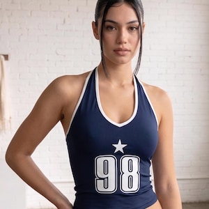 Peut inclure: Haut de style dos nu bleu marine avec bordure blanche, orné d'une étoile blanche et du chiffre "98" en blanc gras. Le haut a une encolure en V et est fait d'un tissu lisse. Un haut élégant et sportif.