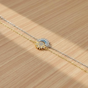 Puede incluir: Dos pulseras delicadas, una dorada y otra plateada, descansan sobre una superficie de madera. Cada pulsera presenta un dije: un sol con piedras transparentes y una luna creciente con piedras azules. Las pulseras están unidas por finas cadenas.