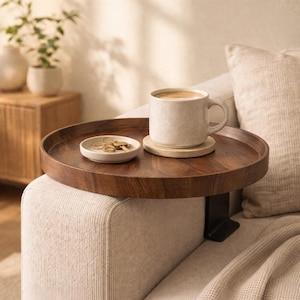 Op de afbeelding: Een ronde, donkerbruine houten sofa tray met een zwarte metalen klem. Een beige mok met koffie staat op een onderzetter en een klein wit schaaltje bevat sleutels. De tray is bevestigd aan een beige bank.