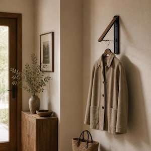 Op de afbeelding: Een aan de muur gemonteerde kapstok met een donkere metalen beugel en een houten arm. Een beige jas hangt aan een houten hanger. Een geweven mand staat eronder op de vloer. De scène speelt zich af in een neutraal getinte entree.