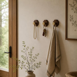 May include: Three wooden wall hooks with brass accents. One hook holds a rope loop, another holds keys, and the third holds a beige towel with a striped border. The hooks are mounted on a light-colored wall.