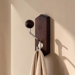 Pode incluir: Um gancho de parede de madeira castanha escura com um gancho de metal prateado e um botão redondo castanho escuro. Um cachecol bege está pendurado no gancho. O gancho está montado numa parede bege clara.