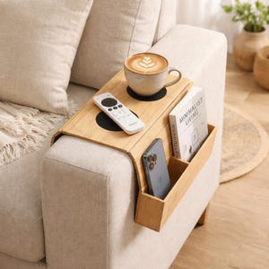 Puede incluir: Una bandeja de brazo de sofá de bambú con un portavasos incorporado, un soporte para control remoto y un bolsillo lateral para un libro y un teléfono inteligente. Una taza de café con arte latte está en la bandeja. La bandeja está en un sofá beige.
