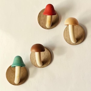 May include: Four wooden mushroom wall hooks in red, tan, brown, and teal. Each mushroom has a white stem and a colored cap, mounted on a round wooden base. The hooks are designed for wall mounting and add a whimsical touch to any room.