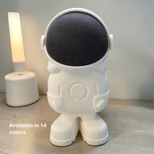 Może przedstawiać: Biały głośnik w kształcie astronauty z ciemnoszarą osłoną głośnika. Głośnik ma okrągłą głowę i korpus z ramionami i nogami. Tekst "Available in 14 colors" znajduje się na dole obrazu.