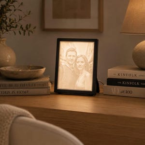 Pode incluir: Uma caixa de luz retangular com moldura preta exibindo um retrato em litofania iluminado em tons quentes de um casal. A imagem apresenta um homem e uma mulher em um estilo monocromático suave e brilhante, dentro de uma moldura simples.