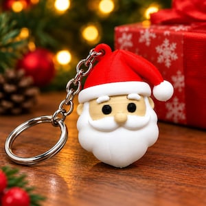 Könnte beinhalten: Ein Weihnachtsmann-Schlüsselanhänger mit roter Mütze und weißem Bommel, weißem Schnurrbart und Bart sowie schwarzen Augen. Die Figur ist mit einer kurzen Kette an einem silberfarbenen Metallring befestigt und besteht aus glattem, geformtem Kunststoff.