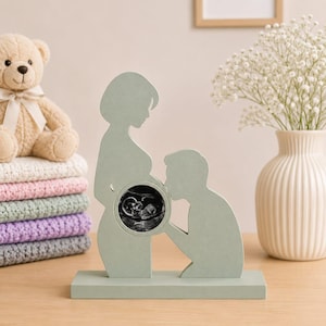 Könnte beinhalten: Ein salbeigrünes Holzornament in Form von zwei Silhouetten, eine stehend und eine kniend, die sich gegenüberstehen. Im Bauchbereich der stehenden Figur befindet sich ein kreisförmiger Ausschnitt mit einem schwarz-weißen Ultraschallbild.