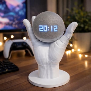 May include: A gray smart speaker with a digital clock display showing 20:12, resting in a white hand-shaped stand. The stand has a circular base. The speaker is on a wooden surface, with a gaming controller and keyboard visible in the background.