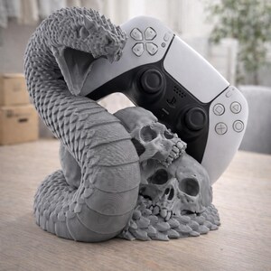 Pode incluir: Suporte cinzento para comando de jogos, impresso em 3D, com uma cobra e caveiras. A cobra enrola-se à volta de duas caveiras, suportando um comando branco e preto. O suporte foi concebido para um comando de jogos.