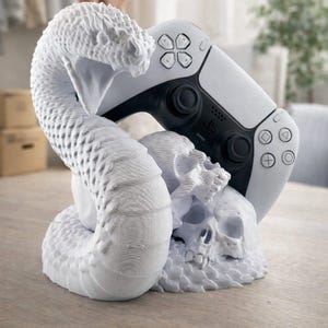 Puede incluir: Una escultura blanca impresa en 3D de una serpiente enroscada alrededor de dos calaveras, sosteniendo un mando de juego blanco y negro. La escultura está sobre una superficie de madera. El mando tiene botones negros y un logotipo de PlayStation.