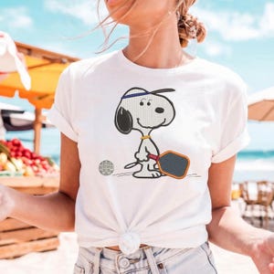 May include: White t-shirt featuring a cartoon Snoopy character playing pickleball. Snoopy is wearing a blue headband and holding a paddle. A pickleball is next to him. The shirt is tied at the waist.