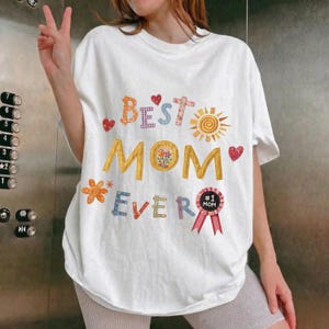 May include: White t-shirt with the text "Best Mom Ever" in colourful, decorative lettering. The design includes hearts, flowers, and a sun graphic. A pink ribbon with "#1 Mom" is also featured.