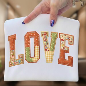 Könnte beinhalten: Weißer Stoff mit dem Wort "LOVE" in Orange, Grün und Stoff mit Blumenmuster appliziert. Die Buchstaben sind mit einer genähten Bordüre umrandet. Das "O" hat ein Tupfenmuster und das "E" ein Blumenmuster.
