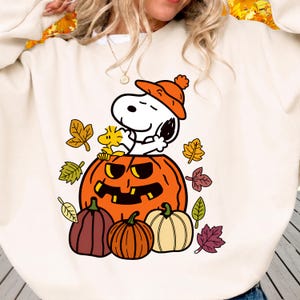 May include: A cream-colored crewneck sweatshirt featuring a graphic print of Snoopy wearing an orange beanie, sitting inside a large orange jack-o'-lantern with Woodstock, surrounded by smaller pumpkins and colorful autumn leaves.