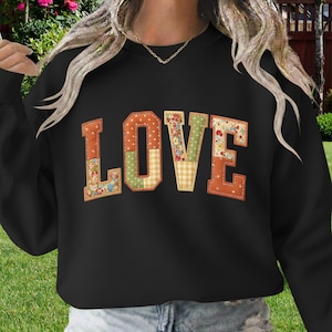 Op de afbeelding: Zwart sweatshirt met het woord "LOVE" in patchwork letters. De letters zijn in oranje, groen en bloempatronen. De sweatshirt heeft een ronde hals.