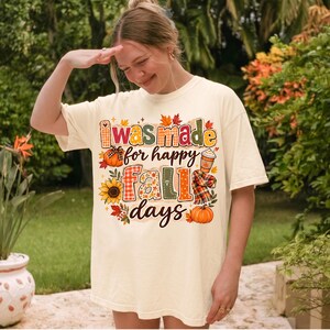 May include: A cream-colored short-sleeve t-shirt featuring a colorful graphic print with the text I was made for happy fall days. The design includes illustrations of pumpkins, a sunflower, a plaid scarf, and a coffee cup with autumn leaves.