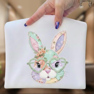 May include: White zippered pouch featuring a colorful bunny design. The bunny has heart-shaped glasses, floral accents, and a patchwork pattern in pastel colors. The pouch is held by a hand.