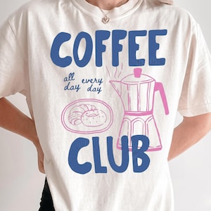 May include: An off-white t-shirt featuring a graphic design with the blue text COFFEE CLUB and the smaller blue text all every day day. The design includes a pink line-art illustration of a moka pot and a plate with a croissant and a donut.
