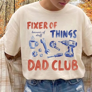 May include: Cream-colored t-shirt with the words "FIXER OF THINGS" in red and blue, along with illustrations of tools. The text "knower of stuff" is above a coffee cup and wrench. The words "DAD CLUB" are at the bottom.