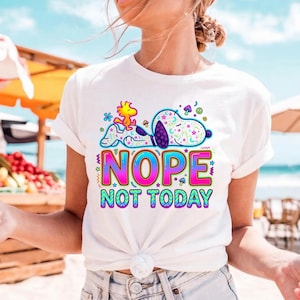 May include: White t-shirt featuring a colorful graphic of Snoopy and Woodstock, with the text "NOPE NOT TODAY" in a vibrant, multi-colored font. The design includes stars, moons, and psychedelic elements.