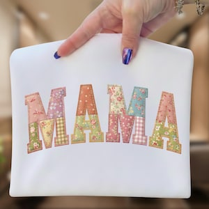 May include: White fabric with the embroidered word "MAMA" in patchwork letters. Each letter features a different floral pattern in shades of pink, green, blue, and yellow. The letters are outlined in a brown stitch.
