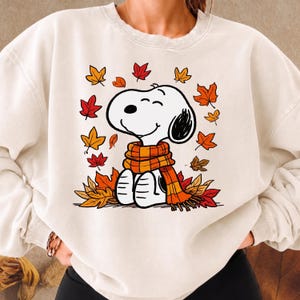 Op de afbeelding: Een crèmekleurige sweater met ronde hals voorzien van een grafische illustratie van het stripfiguur Snoopy met een oranje en gele sjaal met ruitjespatroon, omringd door een verzameling vallende herfstbladeren in rood, oranje en geel.