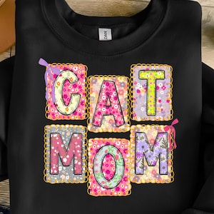 May include: Black sweatshirt with the words "CAT MOM" in colorful, floral-patterned letters. Each letter is in a separate square with a decorative border. The letters are in pink, green, purple, and yellow.