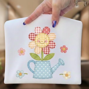 Peut inclure: Pochette blanche avec un motif floral joyeux. La fleur a un visage souriant, des pétales à carreaux et un arrosoir bleu à pois blancs. De petites fleurs entourent le motif principal. La pochette est tenue par une main.