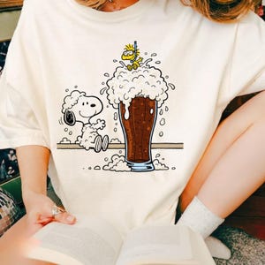 May include: Cream-colored t-shirt featuring a cartoon illustration of Snoopy and Woodstock. Snoopy is sitting next to a large glass of dark beer with a foamy head, while Woodstock is on top of the foam wearing a snorkel.