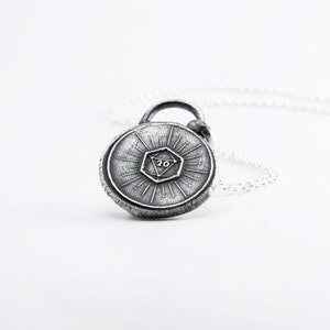 May include: A silver necklace with a round pendant featuring a 20-sided die design. The pendant has a textured surface and a sunburst pattern around the die.