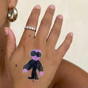 Puede incluir: Una mano con una pequeña pegatina de alienígena morado con gafas de sol negras y un atuendo negro. La mano también tiene un anillo de plata con pequeñas piedras y un pendiente de aro plateado.