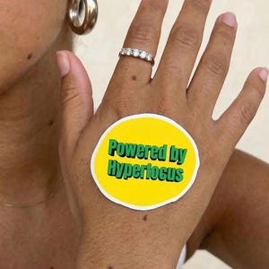 May include: A hand wearing a silver ring with five clear stones and a large silver hoop earring. A yellow sticker with the text "Powered by Hyperfocus" in green is on the hand.