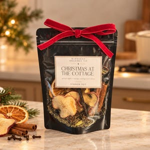 May include: A black pouch containing a simmer pot mix of dried apple slices, orange slices, and cinnamon sticks. The label reads "Midnight Bramble Co. Christmas at the Cottage." A red velvet ribbon is tied in a bow at the top of the pouch.