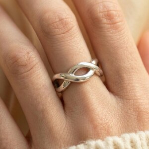 May include: A silver-colored ring with an infinity knot design. The ring is worn on a finger, and the design features two intertwined loops, creating a continuous, flowing shape. The ring is smooth and polished, reflecting light.