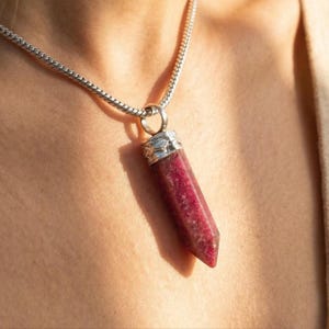 Op de afbeelding: Een zilveren ketting met een hanger met een gefacetteerde, dieprode edelsteen. De hanger heeft een lange, rechthoekige vorm met zilveren accenten aan de bovenkant. De ketting wordt op de huid gedragen.