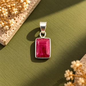 May include: A rectangular pendant featuring a deep red, faceted gemstone set in a polished silver frame with a silver bail at the top. The stone has a rich, saturated color and a smooth, reflective surface, showcasing a classic, clean-cut design.