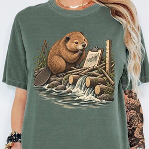 May include: A moss green short-sleeve t-shirt featuring a graphic illustration of a brown beaver sitting on a log dam near a small waterfall. The beaver is holding a clipboard, and the scene includes bulrushes and wooden logs in the water.