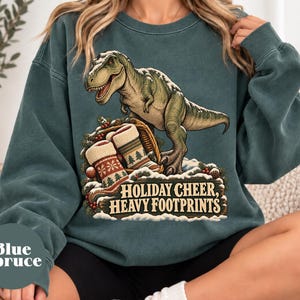 May include: A dark teal crewneck sweatshirt featuring a graphic of a T-Rex standing over a basket filled with two patterned Christmas stockings. The design includes snow, pine branches, and the text "HOLIDAY CHEER, HEAVY FOOTPRINTS."
