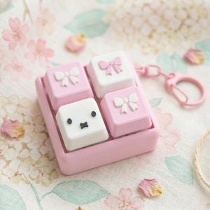 May include: A pink keyboard keycap set with four keys. Two pink keys have white bows, one white key has a cute face, and one white key has a pink bow. The keycaps are in a pink tray with a pink clasp. The background has pink flowers.