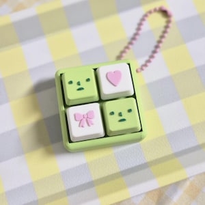 May include: A green square keyring with four smaller squares. Two squares feature cartoon faces, one has a pink heart, and the last has a pink bow. A pink beaded chain is attached.