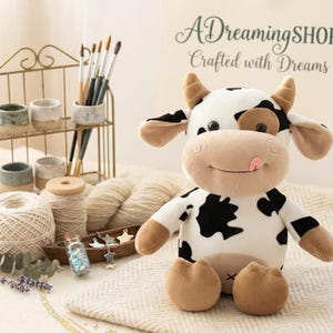 Puede incluir: Un peluche de vaca con manchas negras, cuernos marrones y una lengua rosa. La vaca está sentada sobre una manta color crema. La imagen también incluye materiales de arte, hilo y el texto "ADreamingSHOP: Crafted with Dreams."